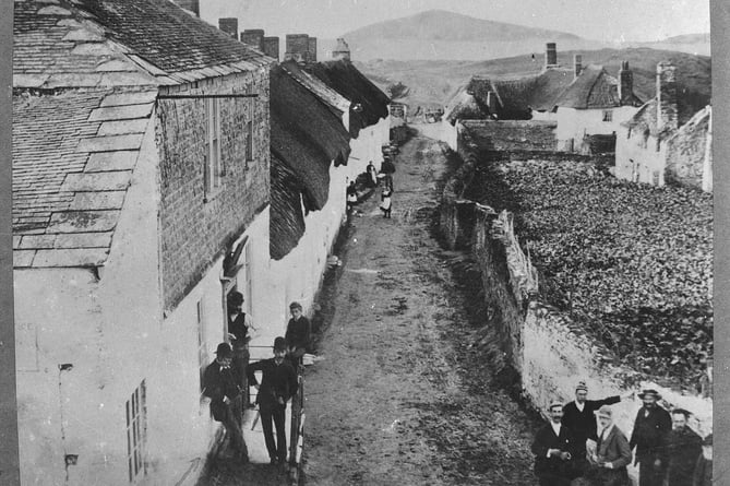 Bantham, looking down the main street. Note Sloop Inn only has two storeys. Various bystanders in front of Sloop, around junction and further down street. Gardens and derelict house on right.