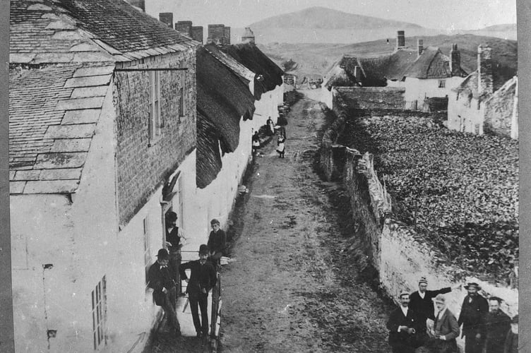 Bantham, looking down the main street. Note Sloop Inn only has two storeys. Various bystanders in front of Sloop, around junction and further down street. Gardens and derelict house on right.